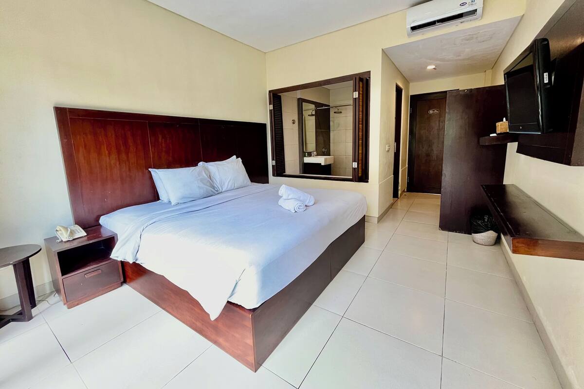Una habitación de hotel moderna con una cama grande, un teléfono en la mesita de noche, un televisor montado en la pared y un baño visible a través de una puerta abierta.
