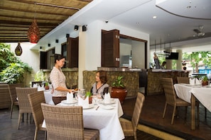 A restaurant with wicker furniture, white tablecloths, and a stone wall backdrop.