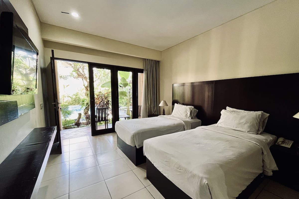 Habitación de hotel con dos camas, televisión y puerta corrediza de vidrio que da a un balcón con vistas a la vegetación.