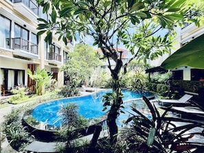 A swimming pool surrounded by lush greenery, with a building featuring balconies and a stone facade in the background. - Suris Boutique Hotel (Kuta)