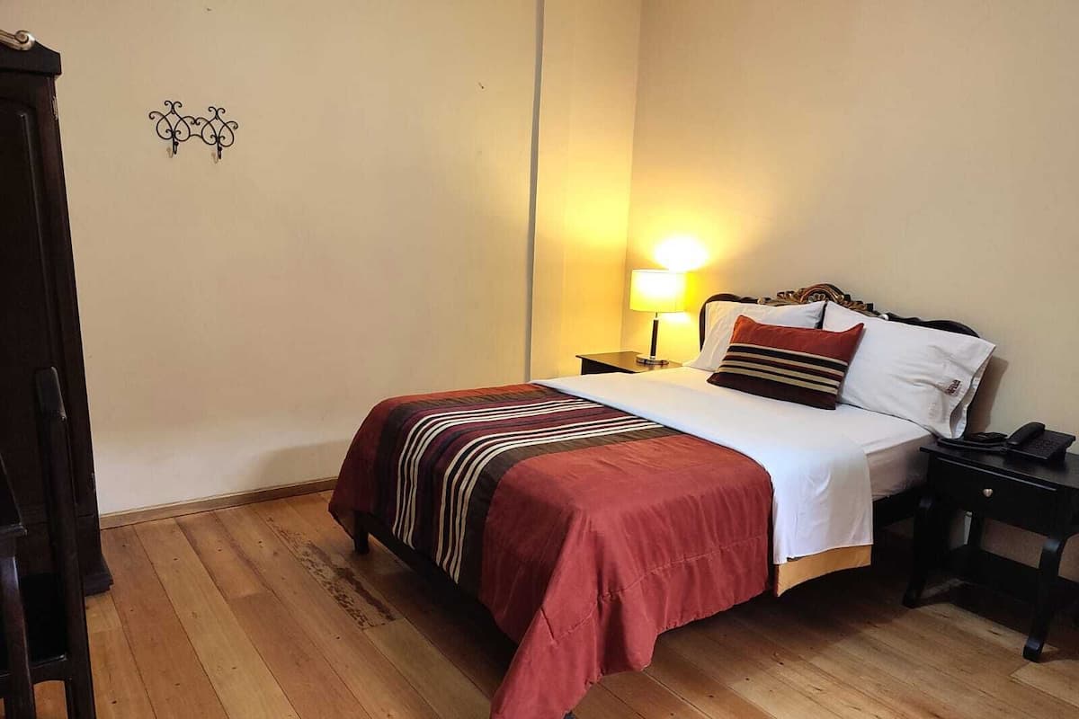 Una habitación de hotel con piso de madera, una cama con colcha a rayas rojas y blancas, una lámpara de noche y una mesita de noche con un teléfono.