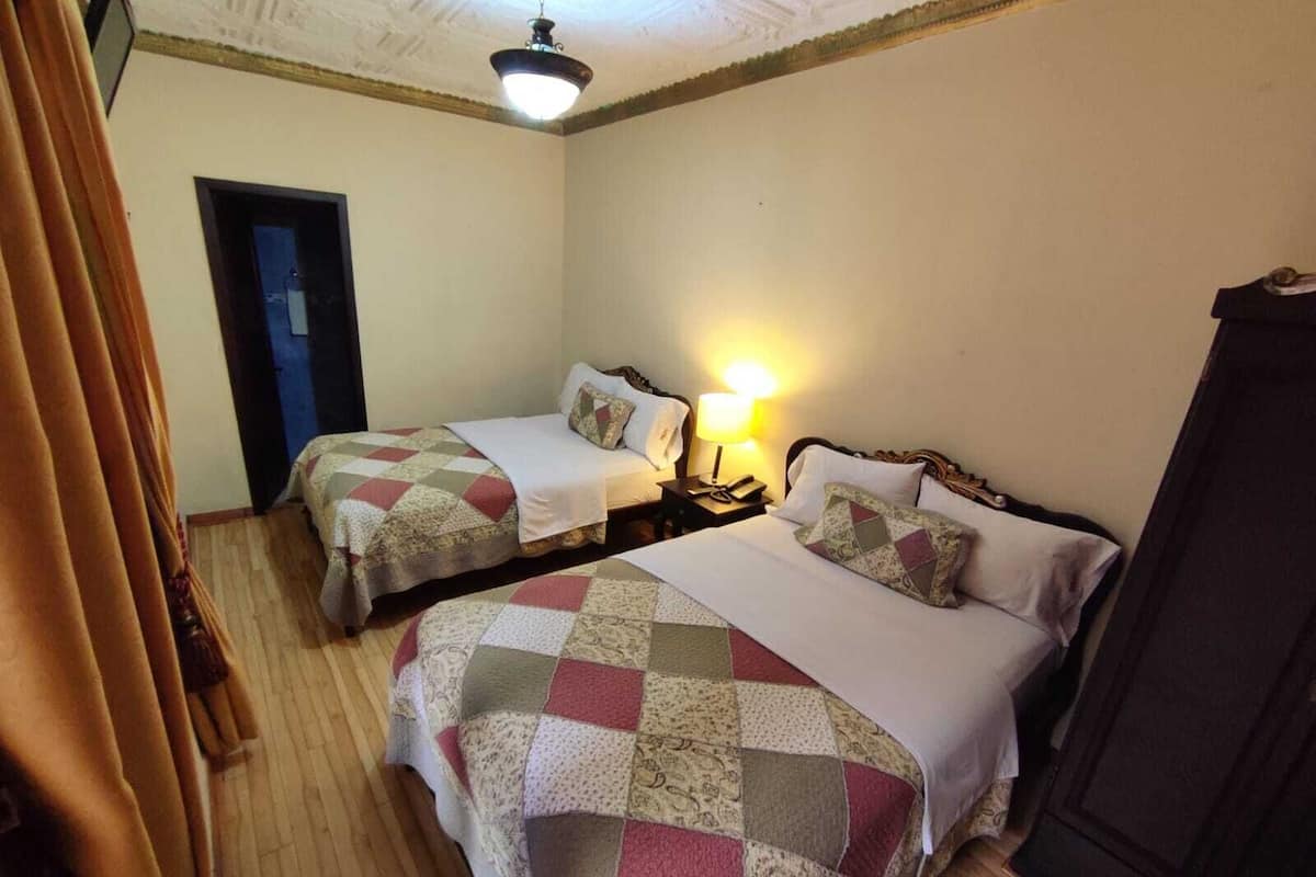 Una habitación con dos camas, cada una con colcha acolchada y almohadas. Entre las camas, un mesita de noche con una lámpara. El suelo es de madera y hay una puerta que da a otra zona.