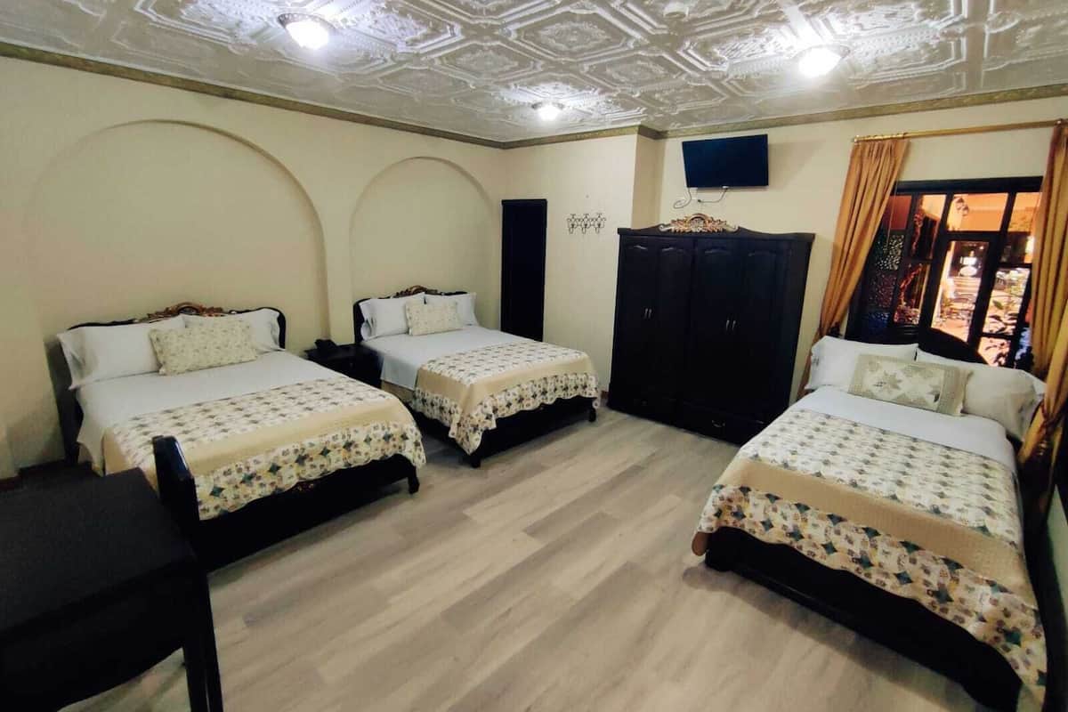 Una habitación con tres camas, un armario de madera oscura, un televisor de pantalla plana montado en la pared y una ventana con cortinas.