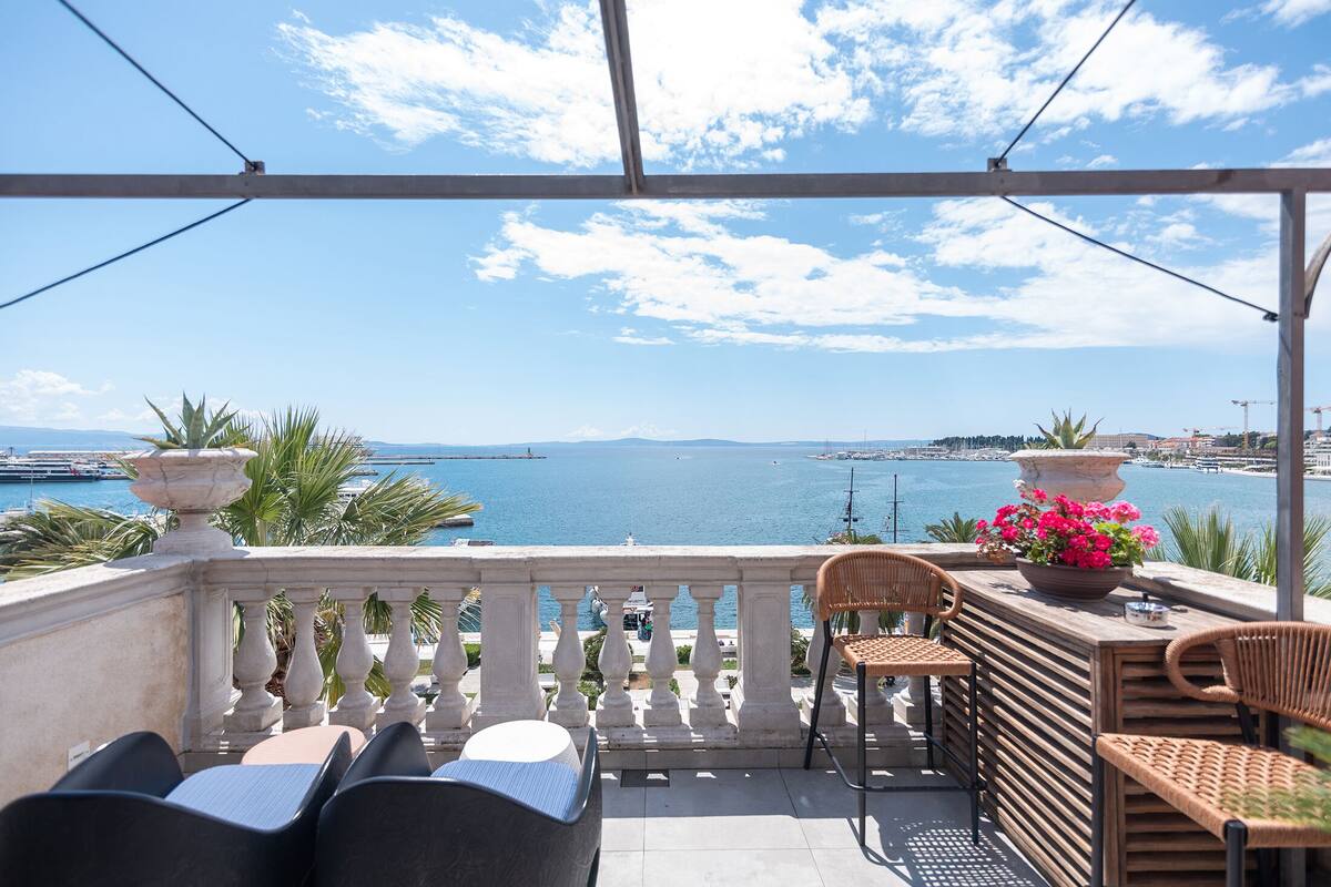 A balcony with a view of the sea, wicker furniture, and a flower vase.
