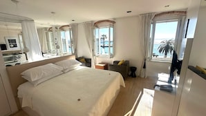 A hotel room with a large bed, a bedside table, a mirror, a window with a view of the sea, and a bathroom with a sink and mirror. - Riva Luxury Suites (Split)