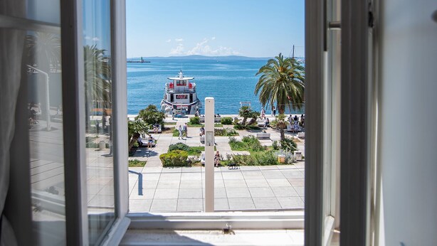 A view from a balcony with a clear sky, palm trees, and a distant building by the sea. - Riva Luxury Suites (Split)