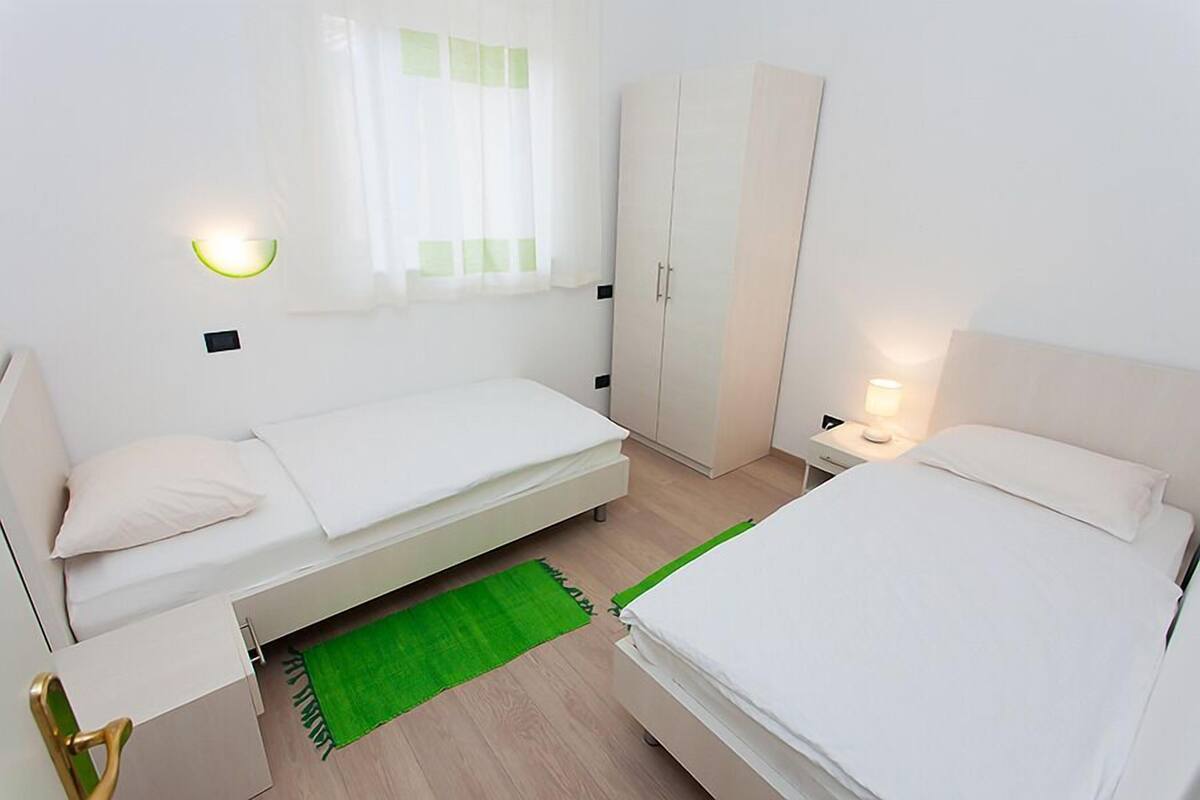 A compact bedroom with two beds, a wardrobe, a bedside table with a lamp, and a green rug.