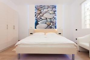 Un dormitorio moderno con una cama, paredes blancas, un ventanal grande y una obra de arte con guijarros colgada en la pared.