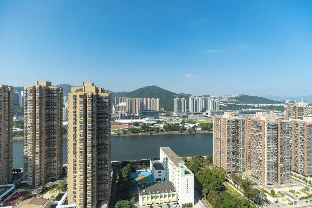 A high-rise residential building complex with multiple apartment buildings, a body of water, and a hilly landscape in the background.