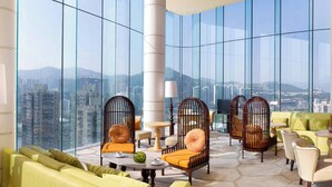 A high-rise hotel room with large windows, a green sofa, a yellow sofa, a round table, and a wooden chair with a cushion.