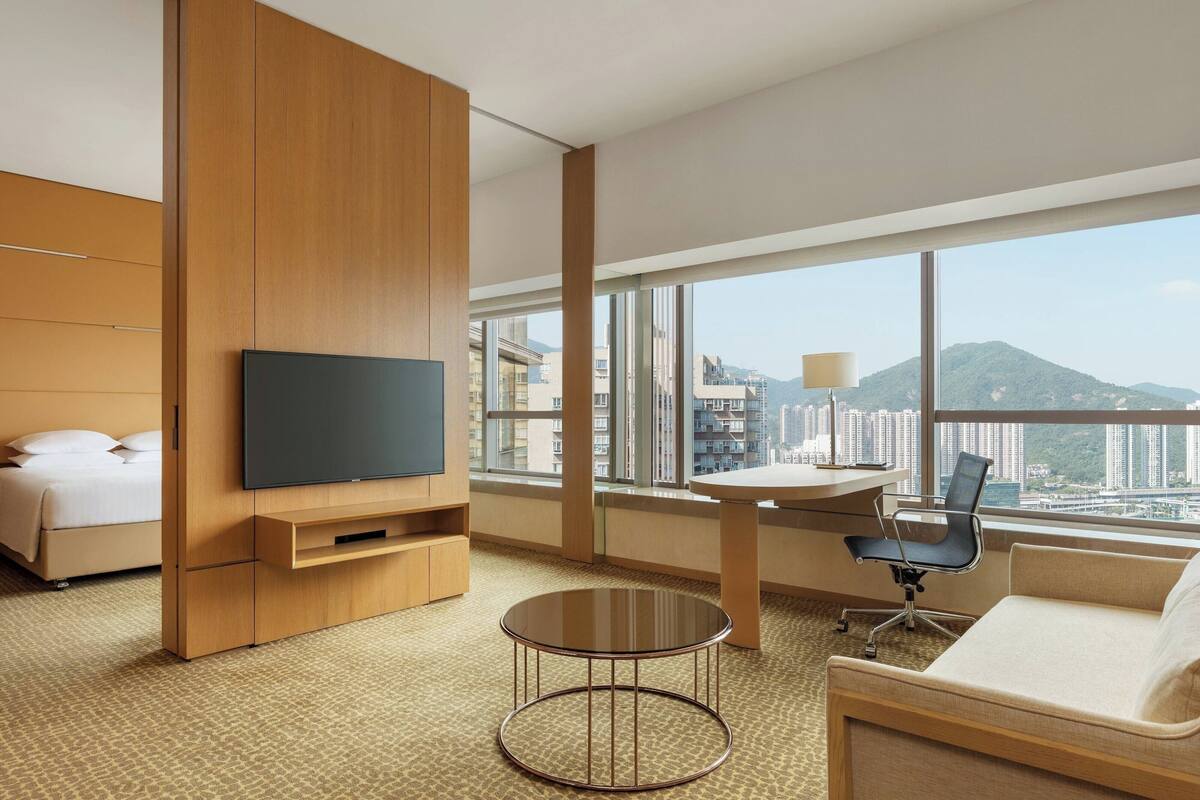 A hotel room with a bed, a TV, a desk with a chair, a round table, and large windows offering a city view.
