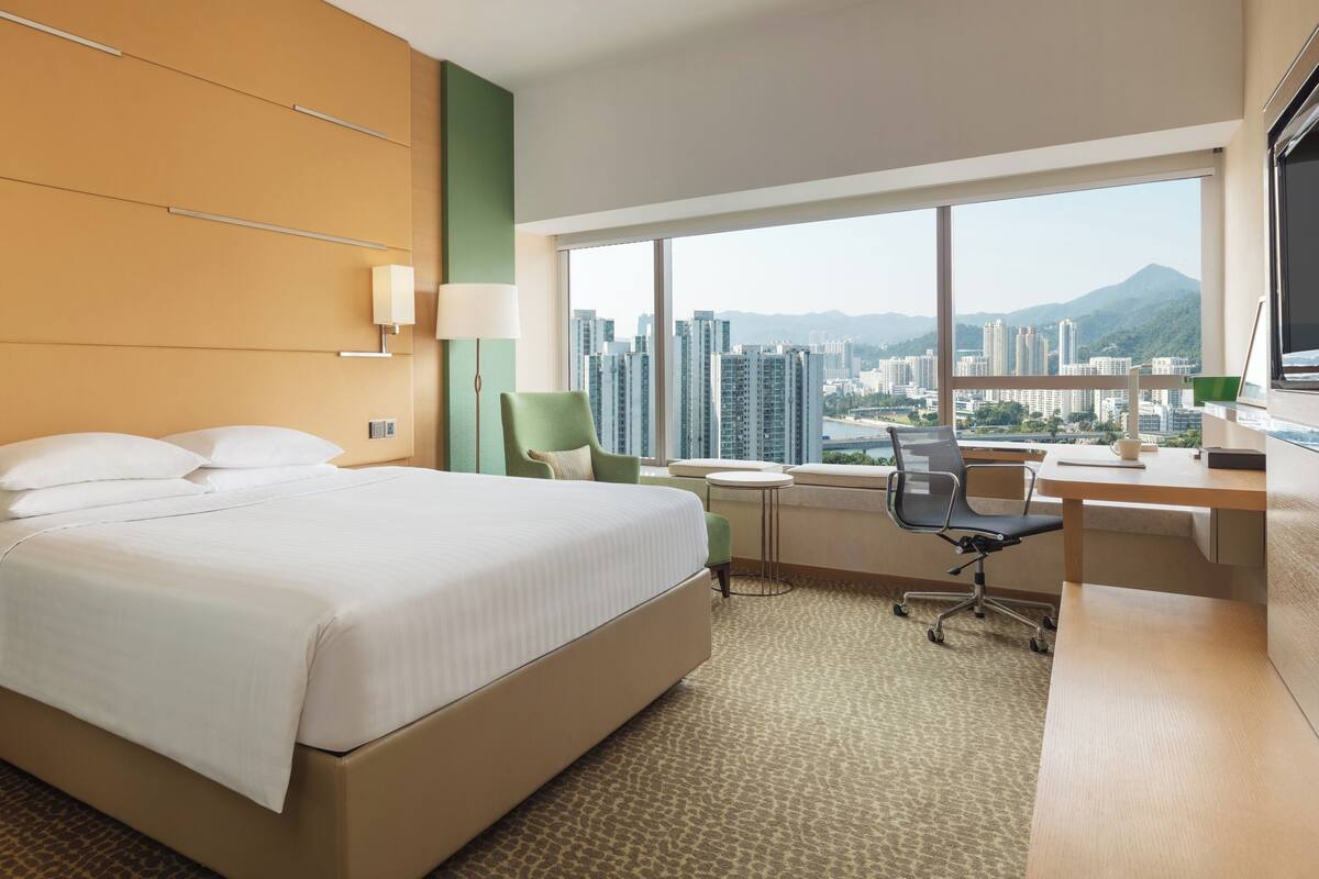 A hotel room with a bed, desk, chair, and large window with a city view.