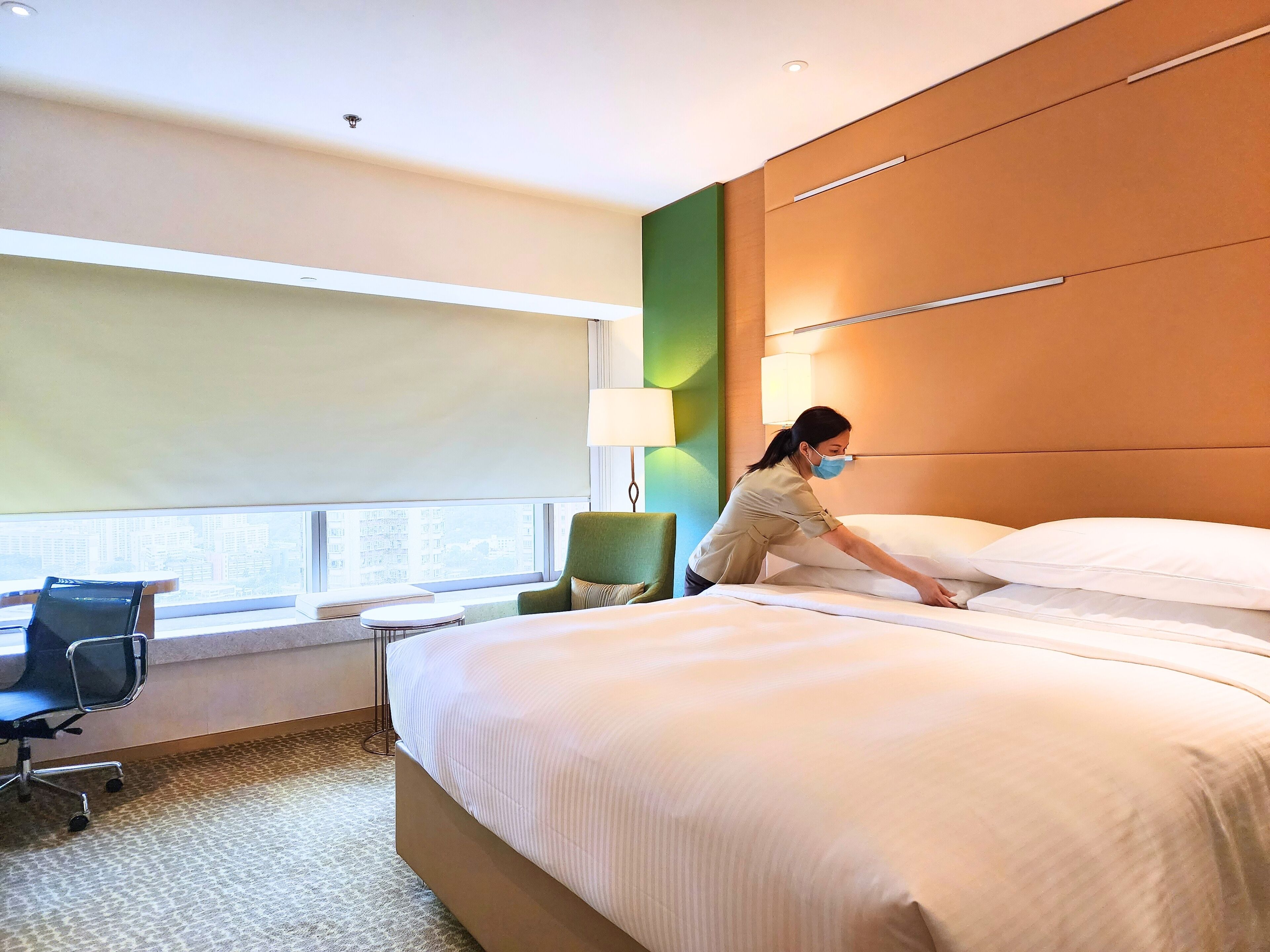 A hotel room with a large bed, a desk with a chair, a window with a view of buildings, and a green accent wall.