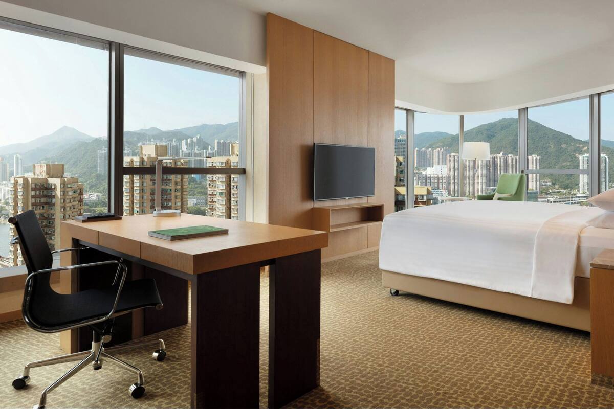 A hotel room with a large bed, a desk with a chair, a television, and a view of a cityscape with mountains.
