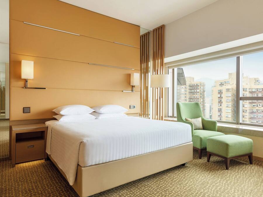 A hotel room with a large bed, a green armchair, a nightstand, a mirror, and a view of the city through large windows.