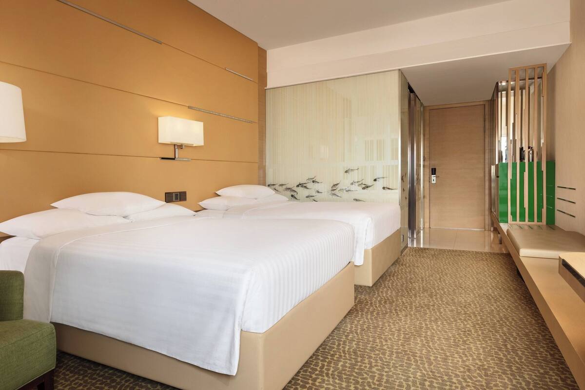 A hotel room with two beds, a green chair, a mirror, and a view of the hallway.