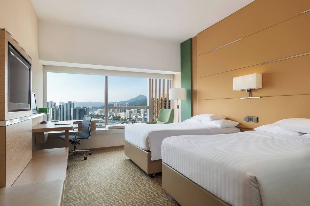 A hotel room with two beds, a desk, a chair, a TV, and a large window offering a city view.