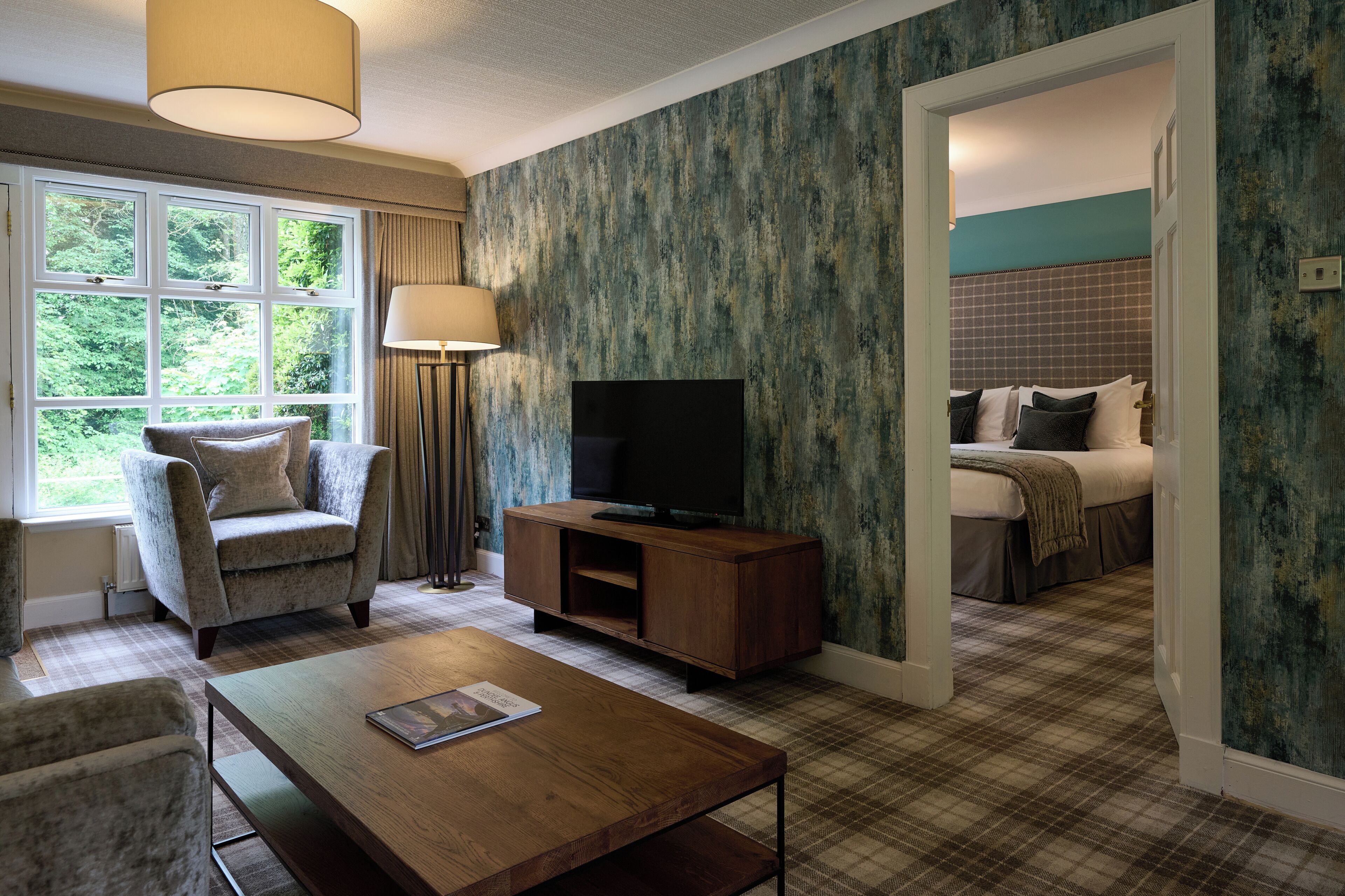 Une chambre d’hôtel avec une grande fenêtre, un téléviseur posé sur un meuble en bois, une moquette à carreaux et un lit recouvert d’une courtepointe à carreaux.