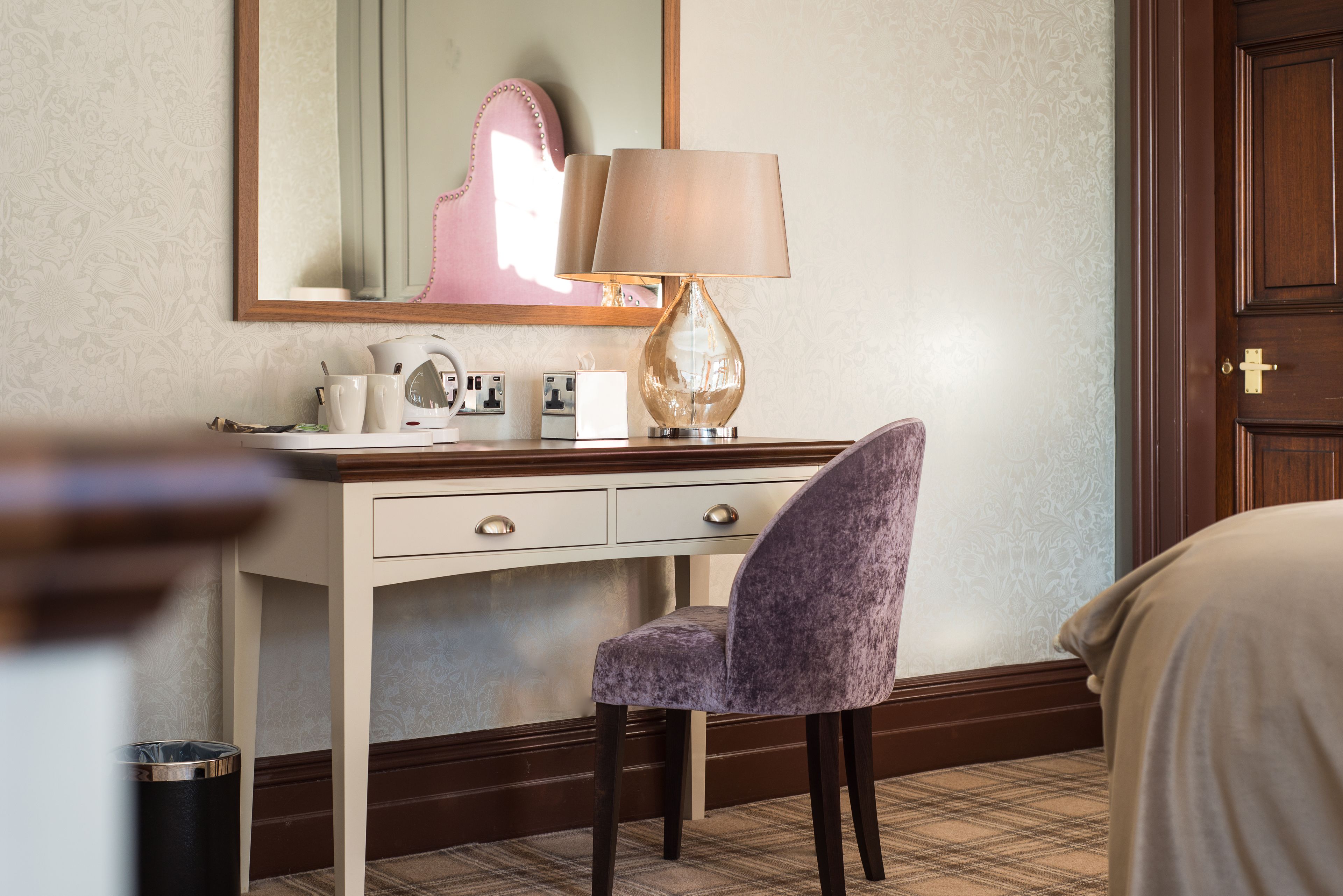 A hotel room with a desk, a lamp, a chair, a bed, a trash can, and a door.