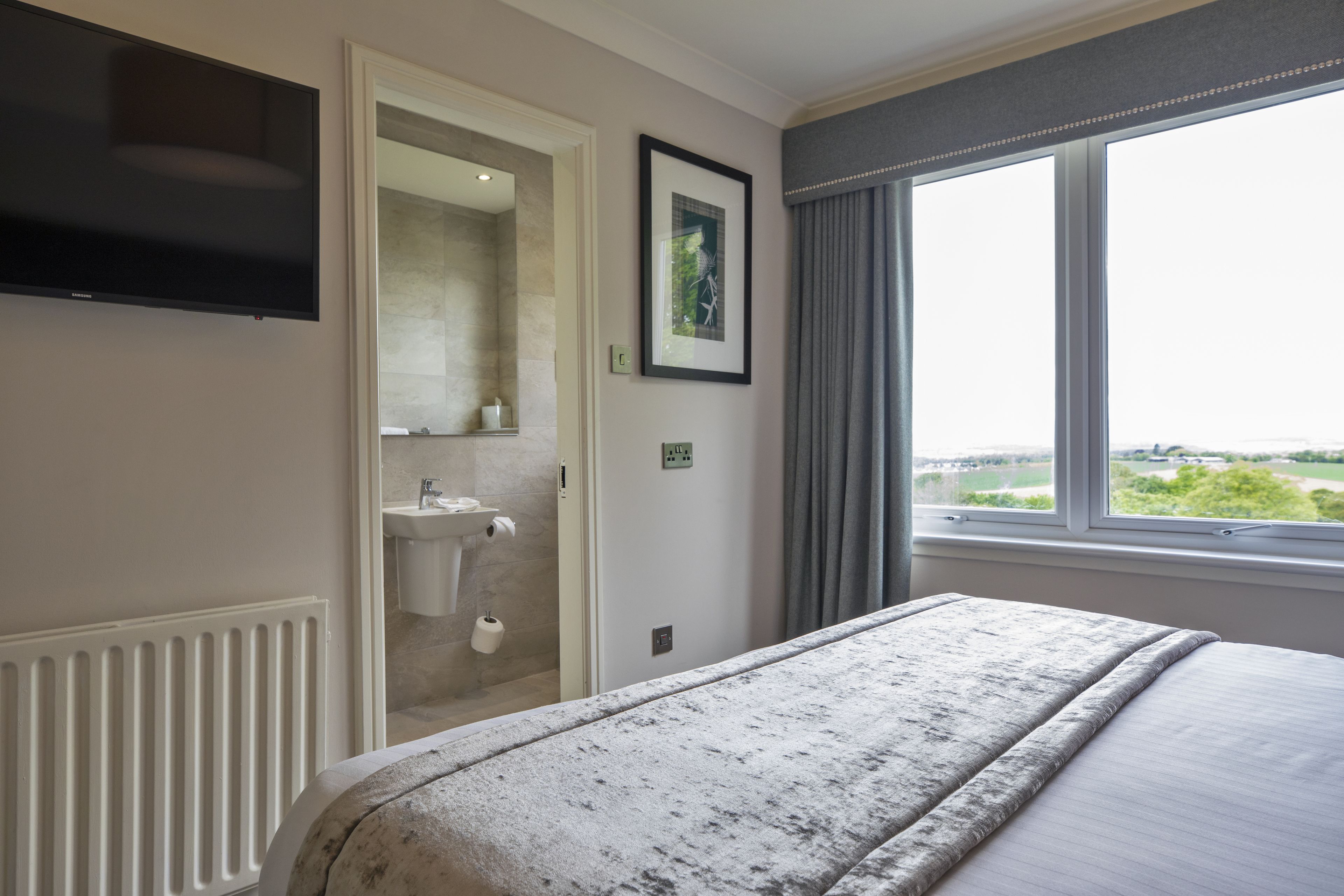 Un dormitorio con una cama grande, un televisor instalado en la pared, una ventana con vistas y un baño visible a través de una puerta abierta.