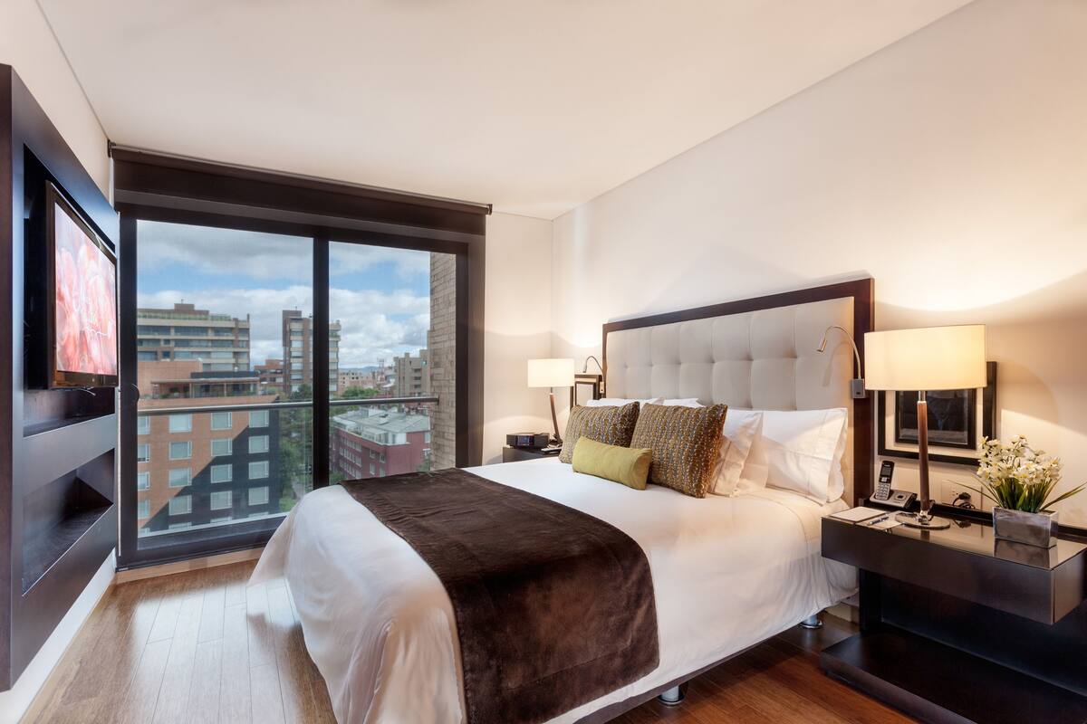 Ein Hotelzimmer mit einem großen Bett, zwei Nachttischen, einem Fernseher und Blick auf die Stadt durch ein großes Fenster.