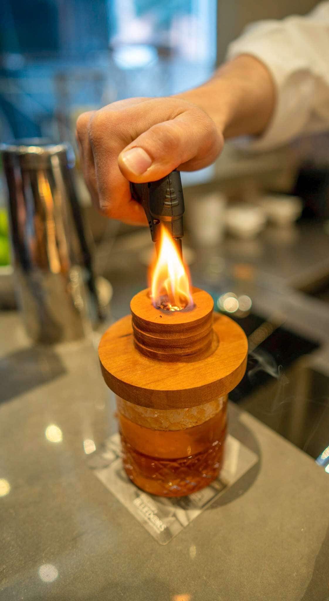 A hand holding a lighter with a flame, positioned over a wooden object on a table.
