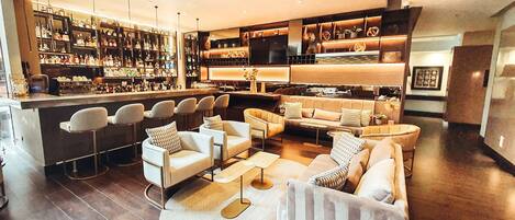 Un bar moderno con diversas opciones de asientos, una estantería repleta de licores y una zona de estar acogedora con sofás y sillones.