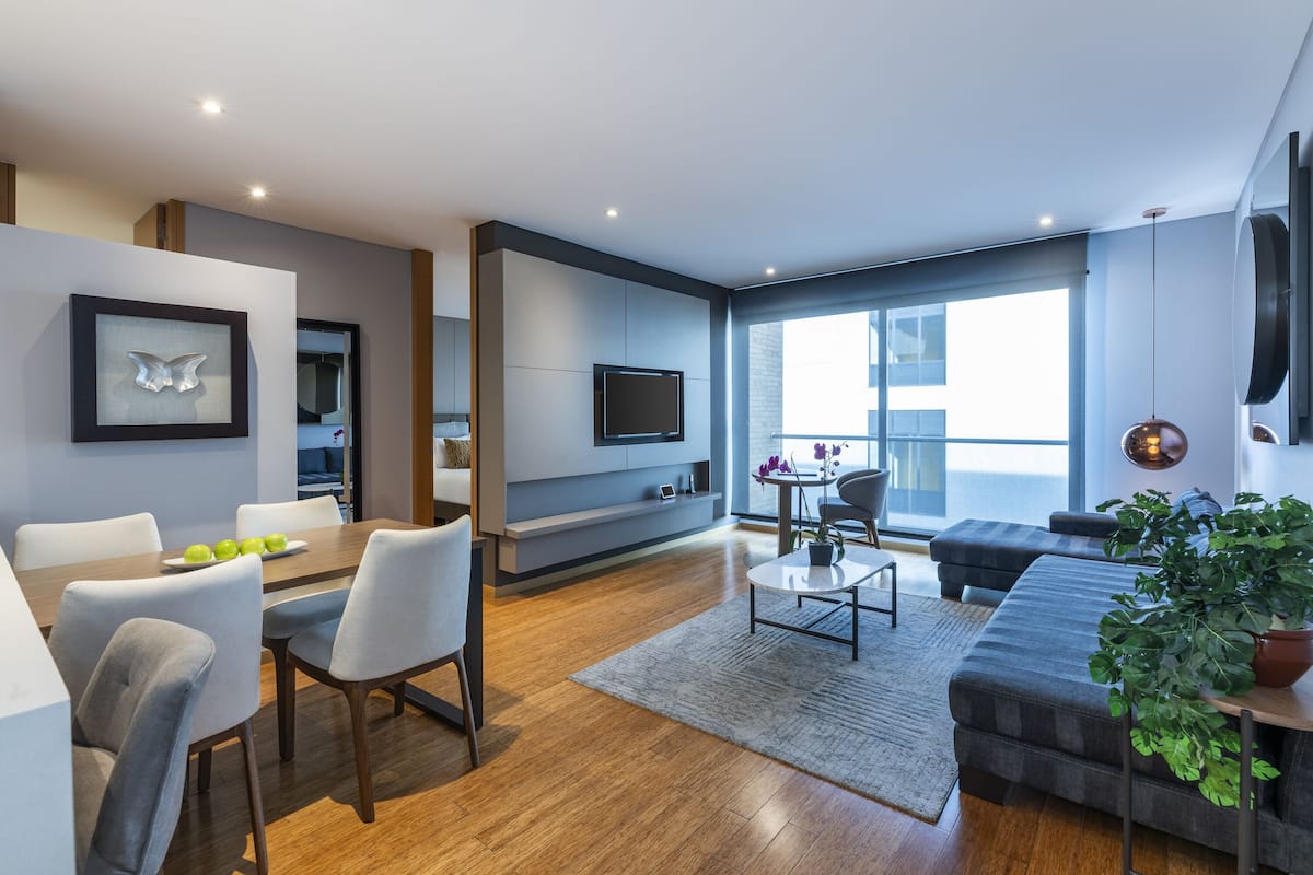 Ein modernes Wohnzimmer mit Flachbildfernseher, grauen Sofas, einem Esstisch mit Stühlen und einem großen Fenster mit Blick auf die Stadt.