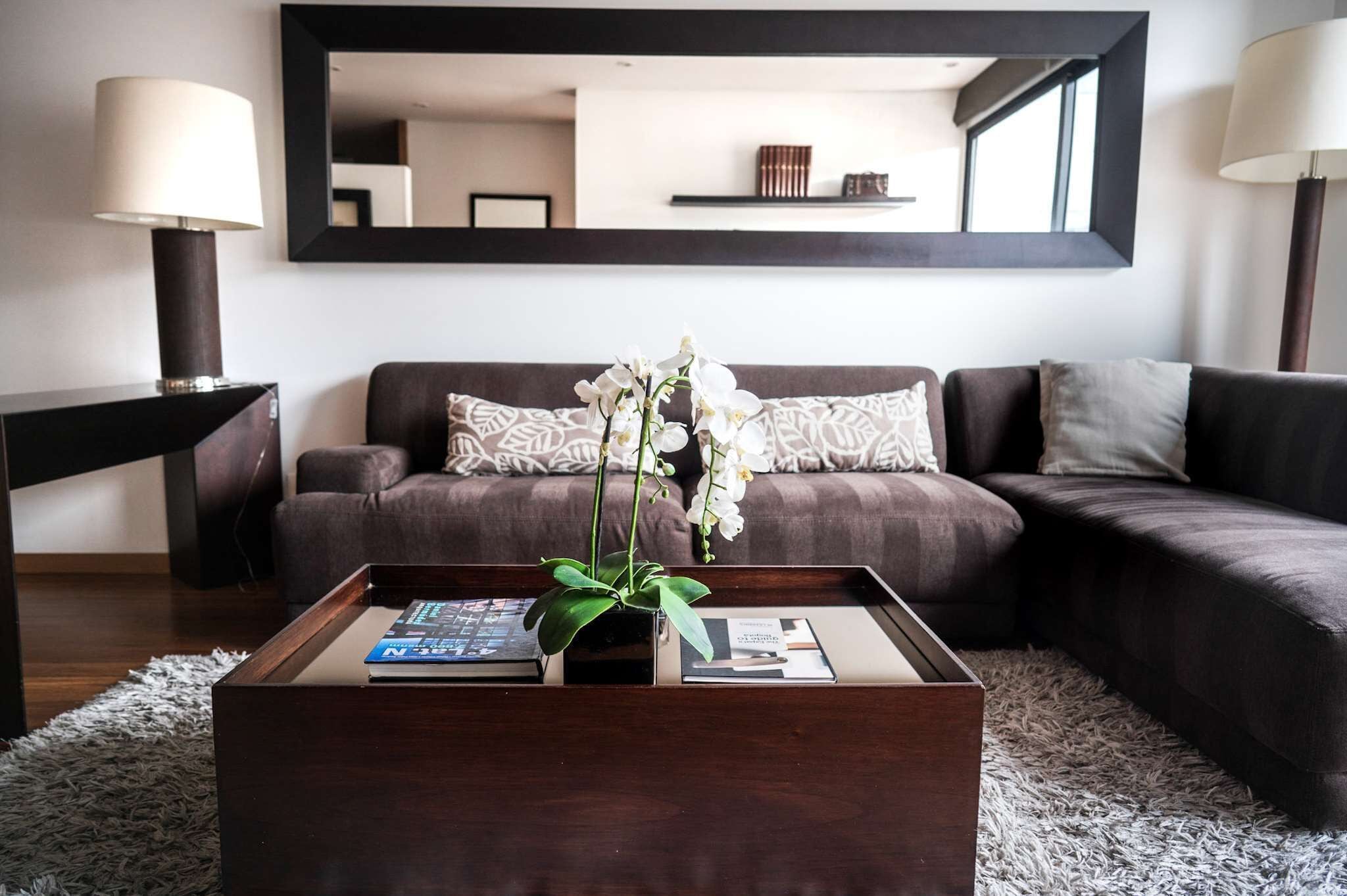 Una sala de estar moderna con un sofá marrón oscuro, una mesa de centro con un jarrón de flores blancas y un espejo grande en la pared.