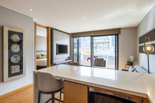 A modern hotel room with a large window, a flat-screen TV, a dining area, and a balcony with flowers.
