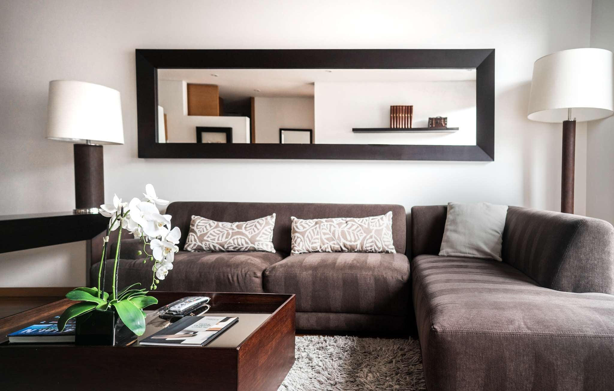 A modern living room with a brown sofa, two patterned pillows, a wooden coffee table, a vase with white flowers, and a large mirror on the wall.