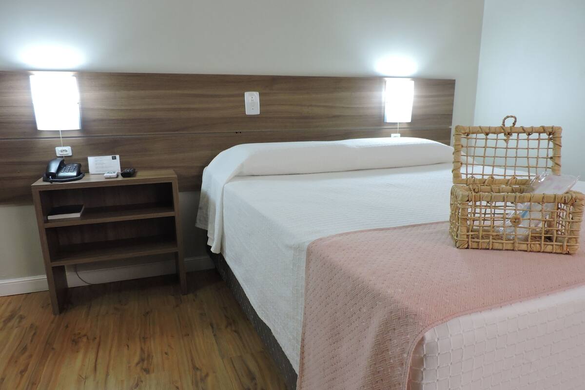 A hotel room with a wooden headboard, a bedside table with a phone and a notepad, a bed with a white comforter, and a wicker basket on the bed.