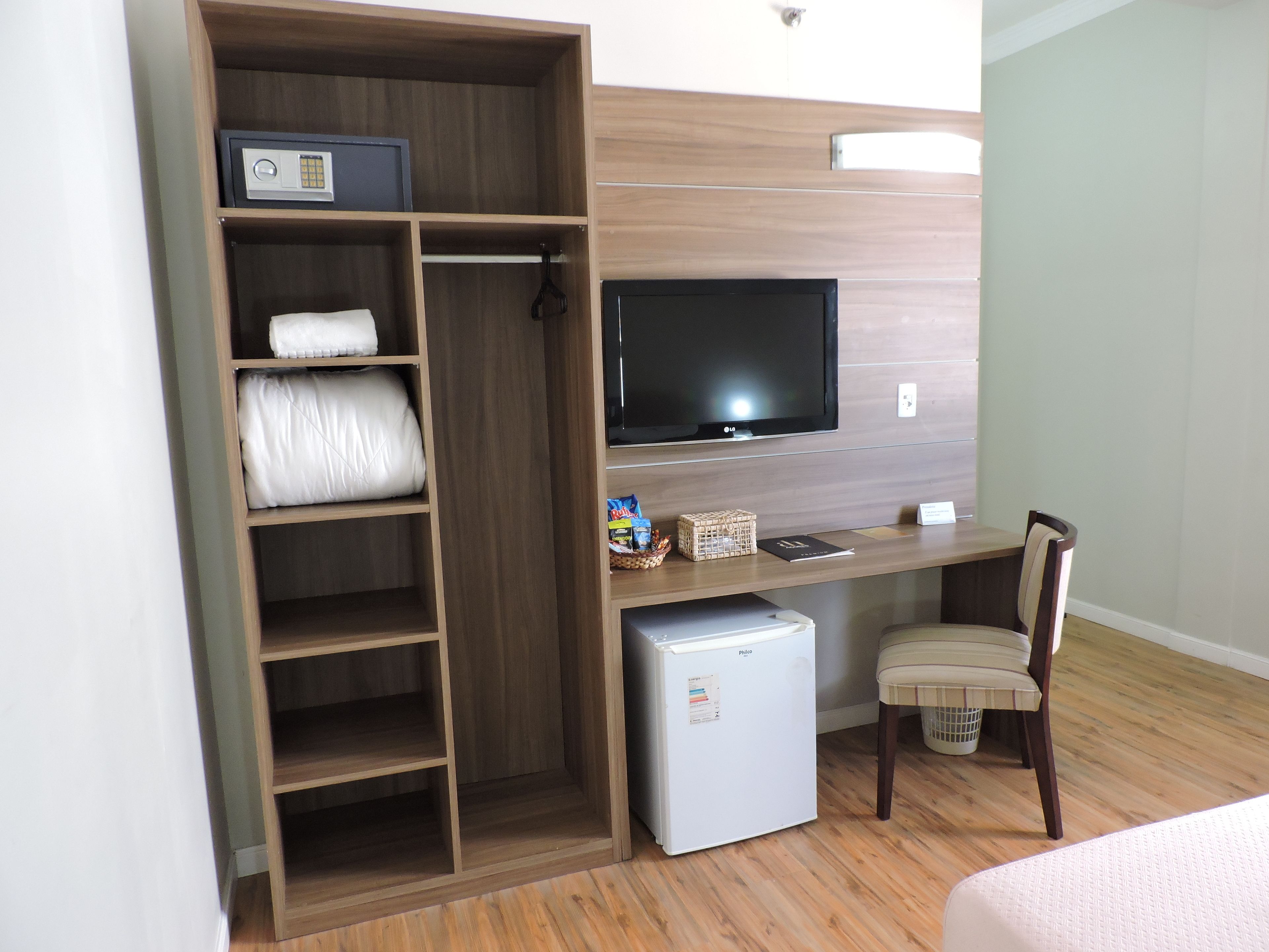 A hotel room with a wooden wardrobe, a small refrigerator, a television mounted on the wall, and a desk with a chair.