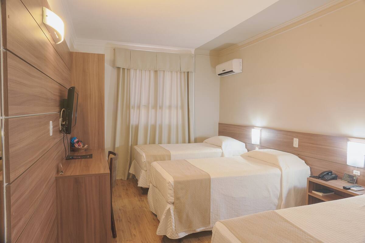 A hotel room with two beds, a wooden desk, a chair, a small table with a phone and a notepad, and an air conditioning unit on the wall.