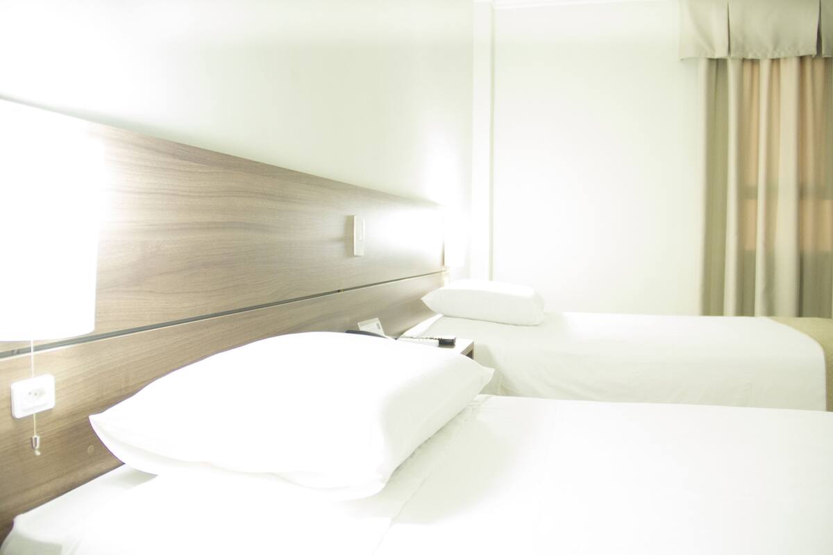 A hotel room with two beds, a wooden headboard, a bedside table, and a window with curtains.