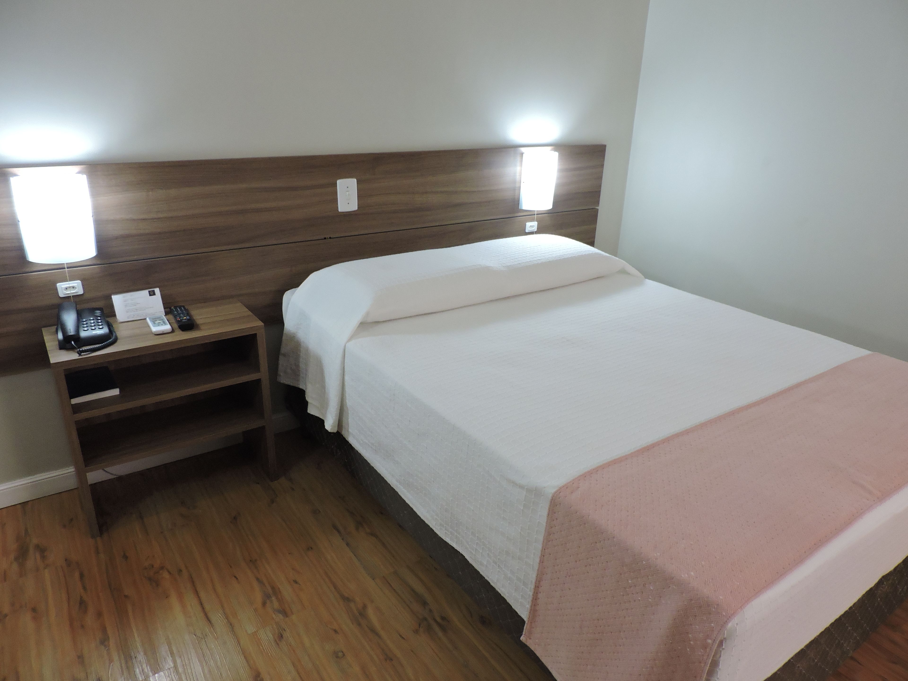 Une chambre d’hôtel avec une tête de lit en bois, une table de chevet avec un téléphone et un bloc-notes, un lit recouvert d’une couvre-lit blanche et rose, et un plancher en bois.