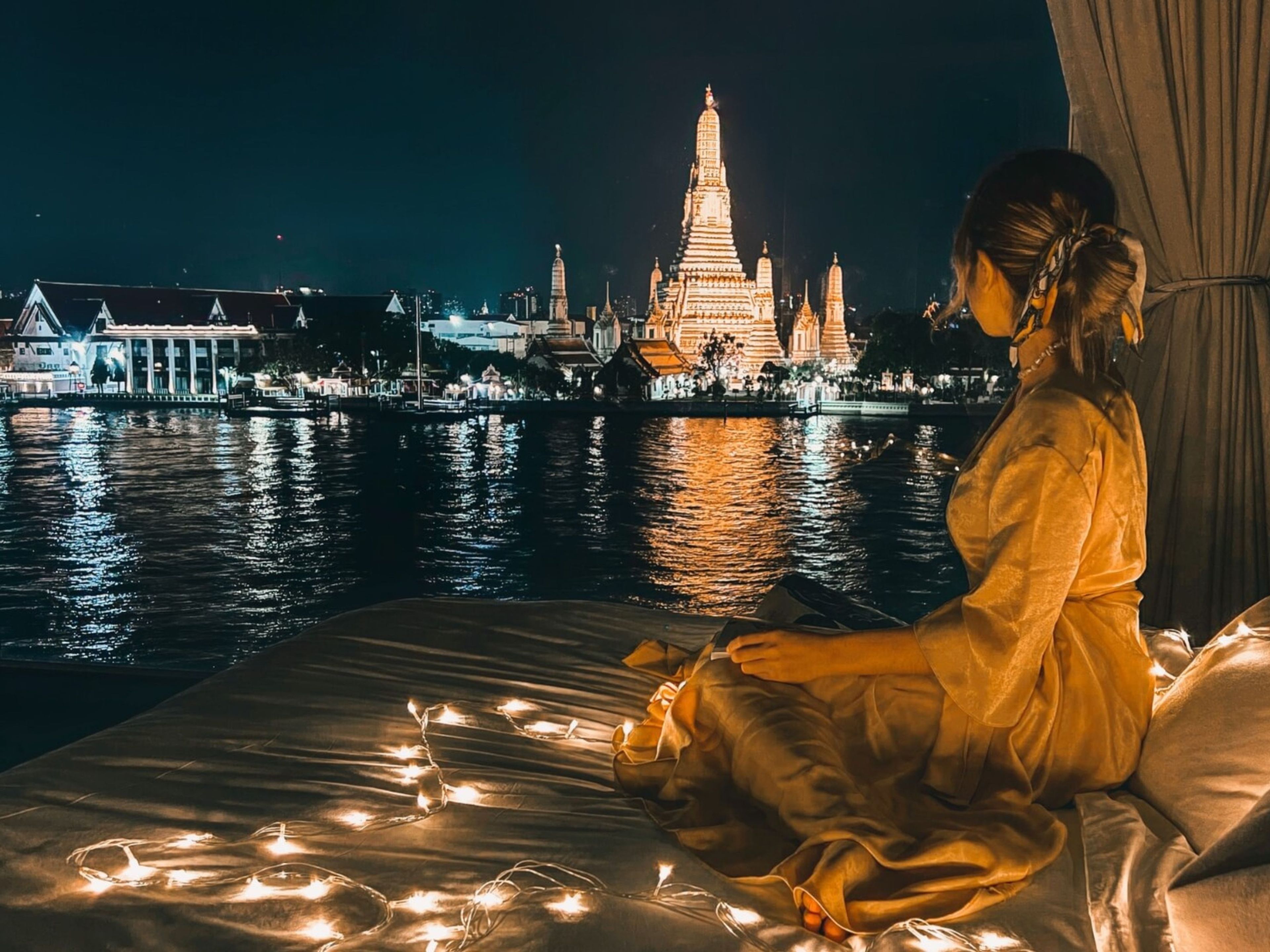Una persona sentada junto a una ventana con vista a una masa de agua y edificios iluminados al fondo.