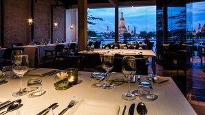 An indoor dining area with tables set for a meal, large windows offering a view of a distant cityscape at dusk. - Sala Rattanakosin Bangkok (Bangkok)