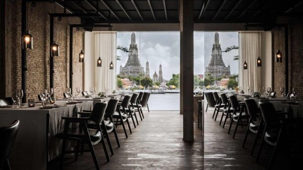 Un restaurante con vistas a un templo. Las mesas están dispuestas con sillas, y la sala tiene una pared de ladrillo rústico y luces colgantes.