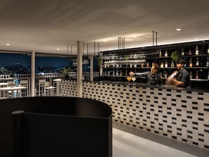 Espaço de bar com padrão quadriculado na parede, balcão e vista noturna da cidade.