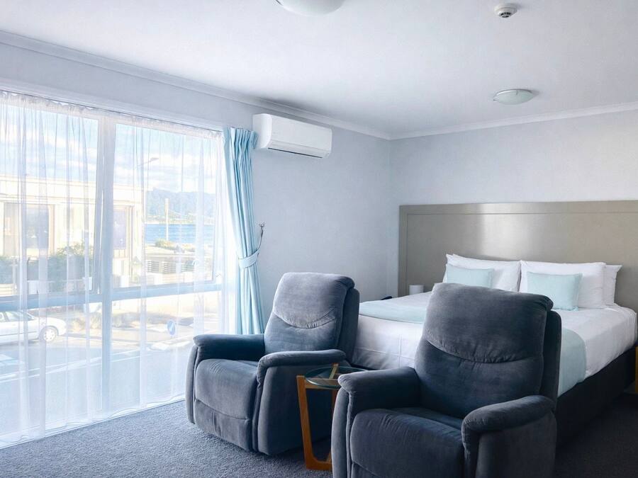 Hotellihuone, jossa on sänky, kaksi nojatuolia, yöpöytä ja suuri ikkuna, jossa on verhot.