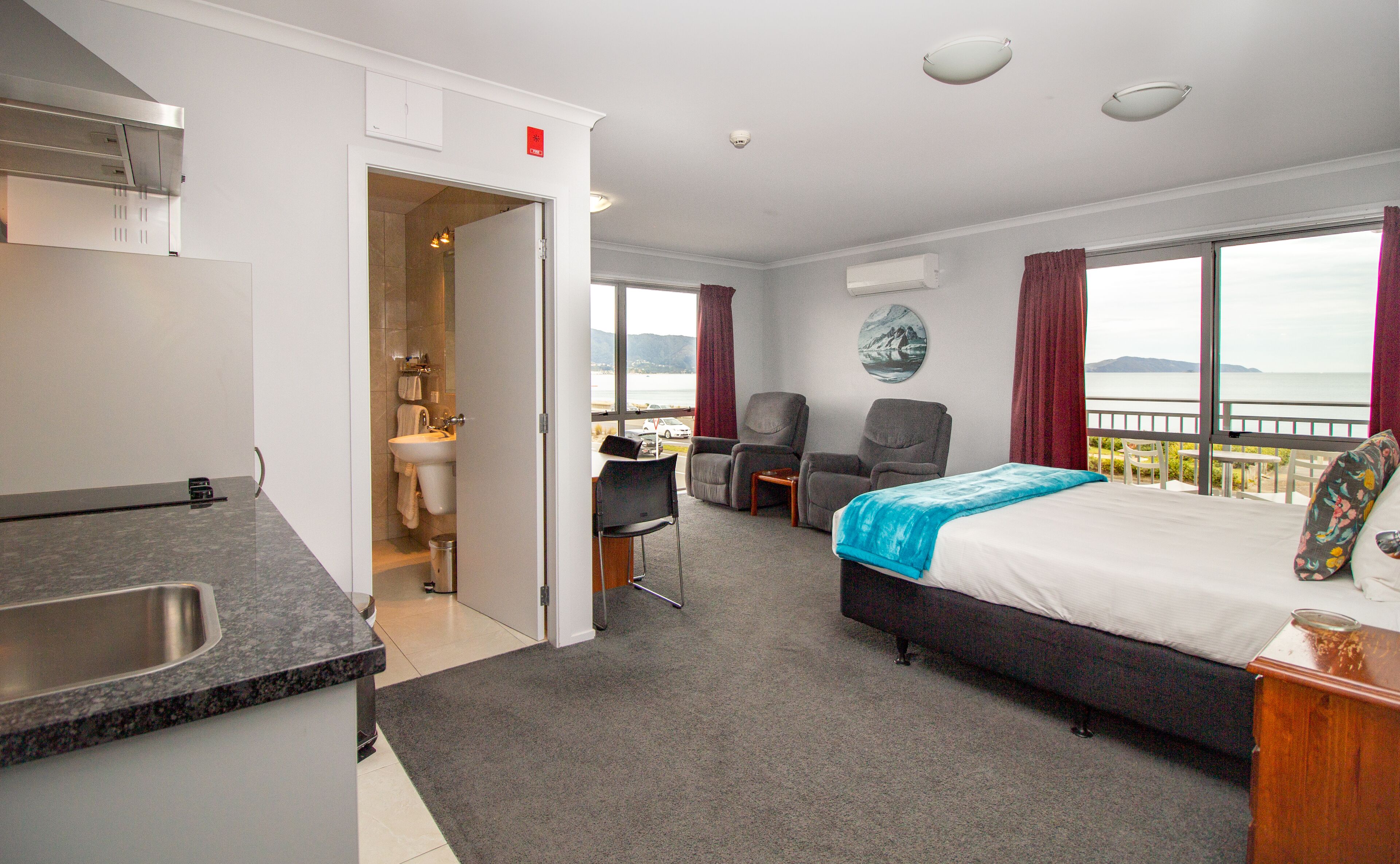A hotel room with a bed, a desk, a chair, a small table, a sink, and a view of the sea.