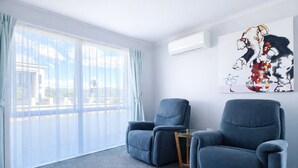 Ein Zimmer mit zwei blauen Sesseln, einem kleinen Glastisch und einem großen Fenster mit durchsichtigen Vorhängen.