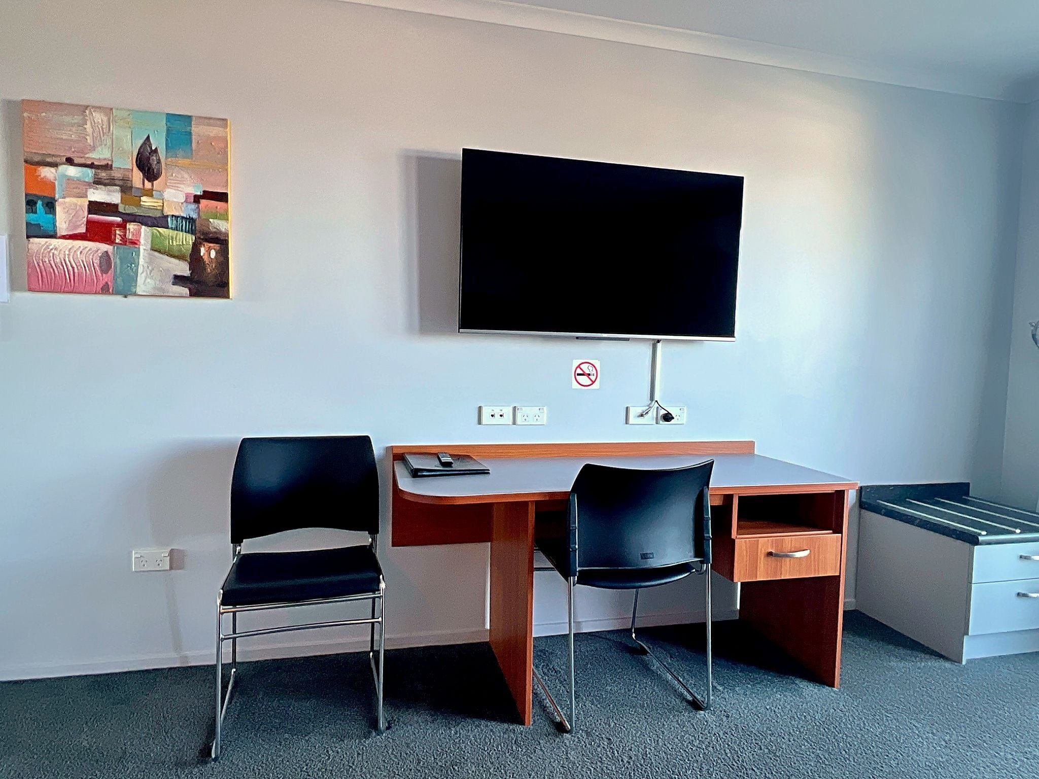 A hotel room with a desk, two chairs, a television, and a painting on the wall.