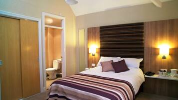 Habitación de hotel con cama, mesita de noche, lámpara y baño con inodoro y ducha.