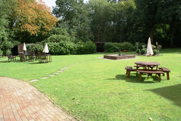 Un jardín bien cuidado con mesas de picnic y sombrillas, rodeado de abundante vegetación y árboles.