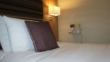 A hotel room with a bed, pillows, a nightstand with a lamp, and a tea dispenser.