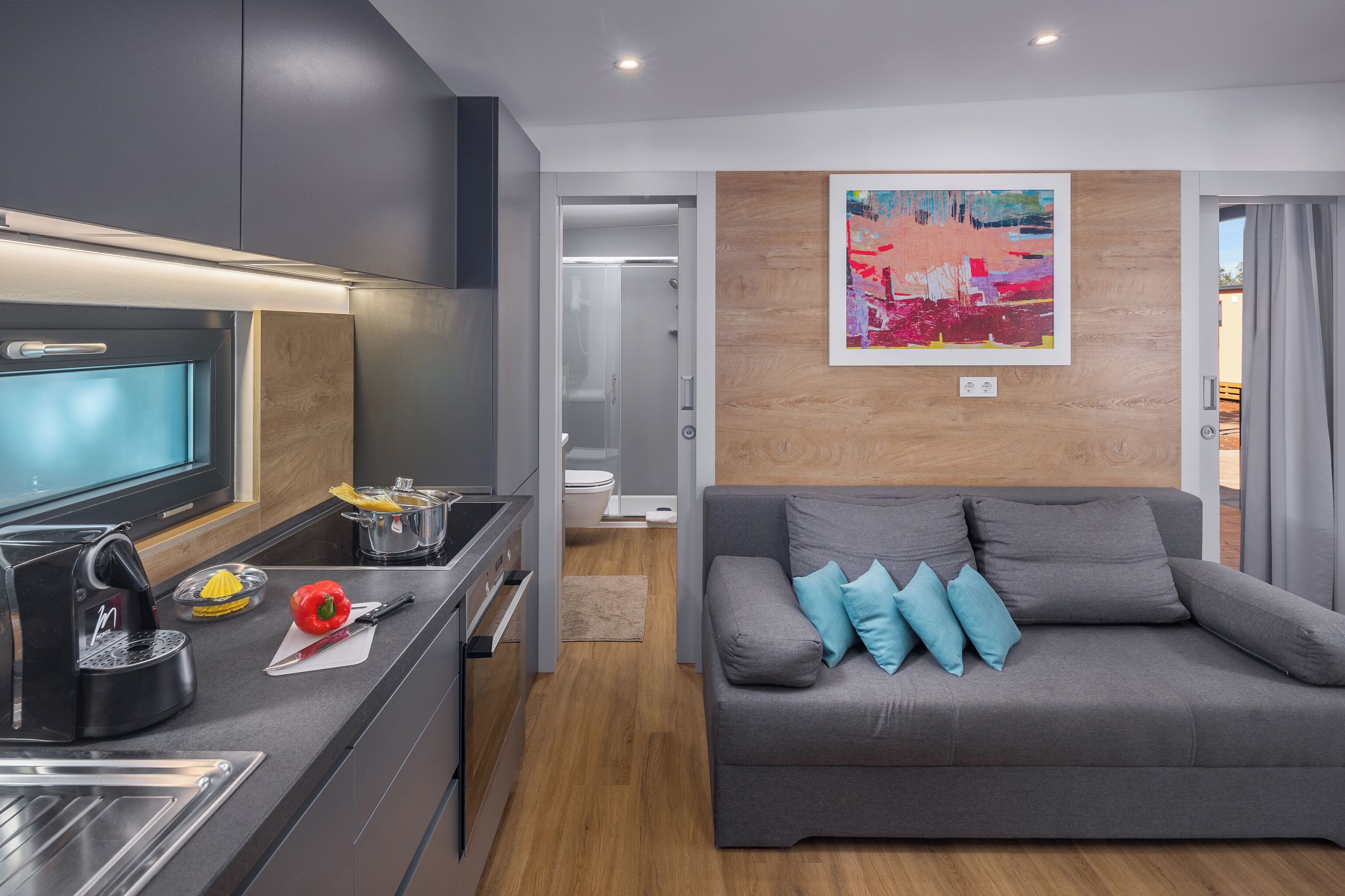 A modern kitchen with a built-in oven, a coffee machine, and a cutting board with a red bell pepper. There is a grey sofa with blue cushions and a wooden accent wall with a colorful painting.