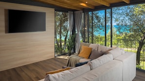 Uma sala de estar moderna com uma grande TV de tela plana na parede, um sofá bege com almofadas, e vista para o mar através de grandes janelas.