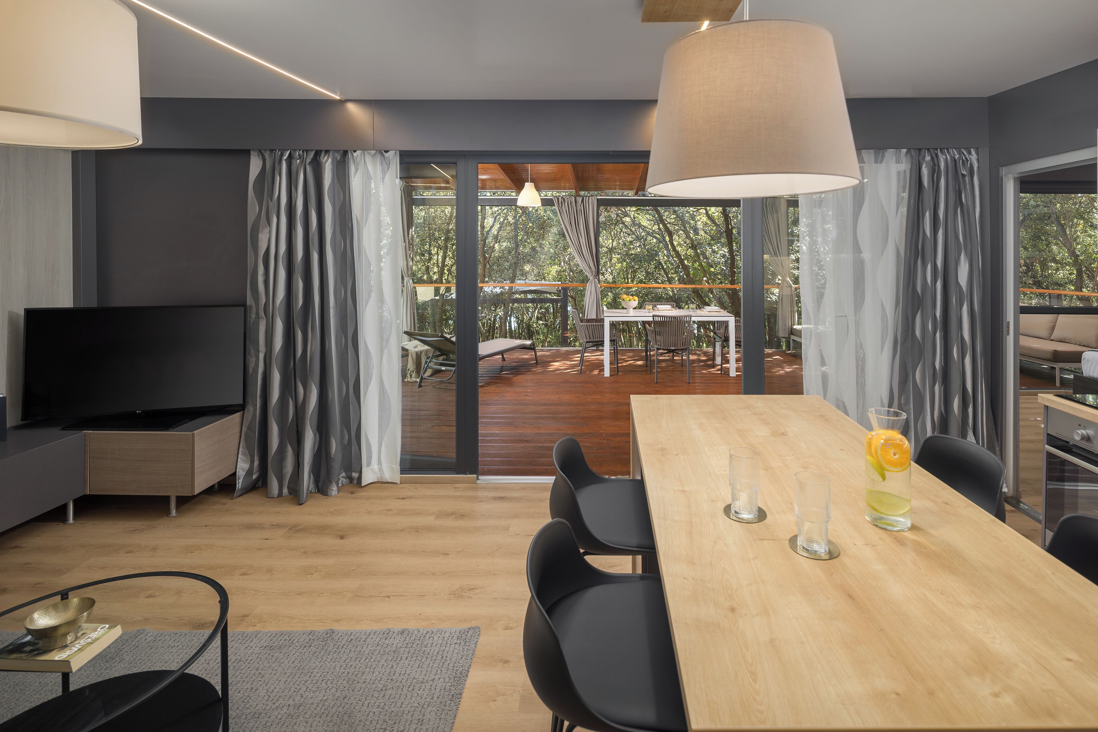 Un salon moderne avec une grande table à manger en bois, des chaises noires, un téléviseur à écran plat et une grande fenêtre avec des rideaux.
